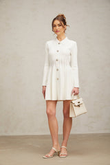 Young woman wearing a white pleated knit mini dress with short sleeves and a fitted waist standing against a plain background