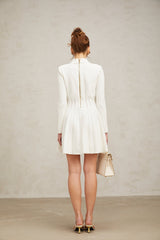 Young woman wearing a white pleated knit mini dress with short sleeves standing against a plain background