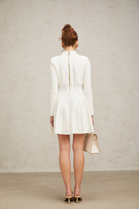Young woman wearing a white pleated knit mini dress with short sleeves standing against a plain background