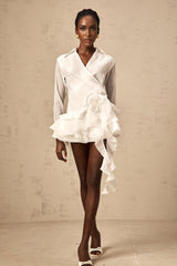 Young woman wearing a white semi sheer mini dress with floral pattern and ruffled details standing against a plain background