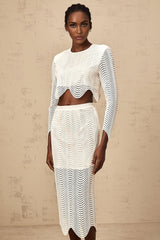 Young woman wearing a white twopiece outfit consisting of a wavy woven top and matching skirt standing against a plain background