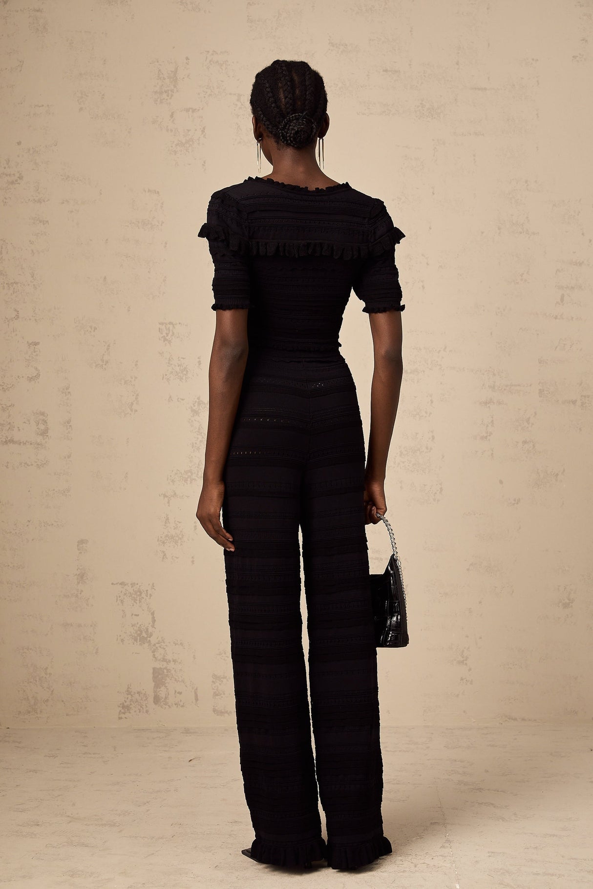Young woman wearing black knitted long sleeve top with ruffled details paired with matching black trousers standing against plain background