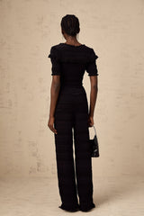 Young woman wearing black knitted long sleeve top with ruffled details paired with matching black trousers standing against plain background