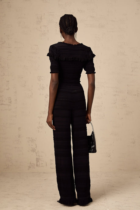 Young woman wearing black knitted long sleeve top with ruffled details paired with matching black trousers standing against plain background