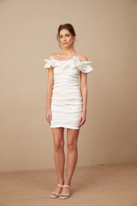 Young woman wearing white offshoulder ruffled mini dress standing against plain background